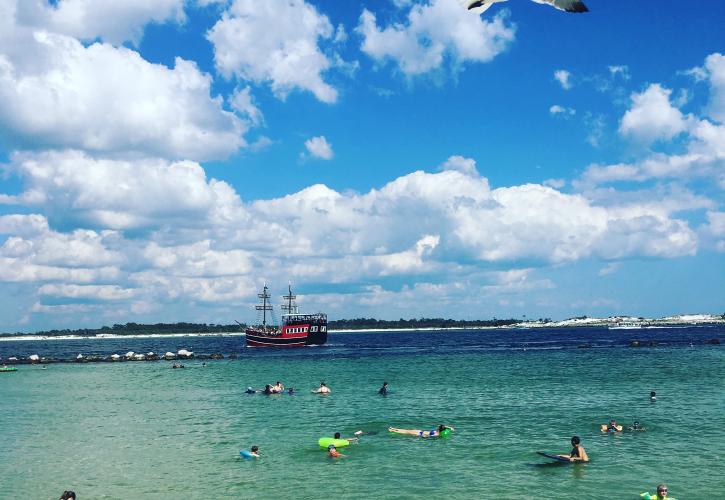 Sea gulls fly overhead and pirate ship sails in the distance while swimmers enjoy emerald green waters.